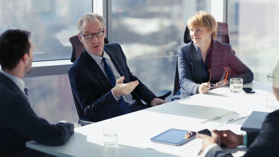 A company's board of directors, with two men talking and a woman looking on and smiling around a board table