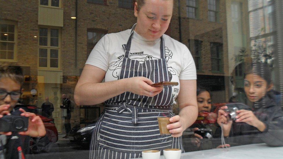 A picture of the owner of cafe taking a picture of her coffee whilst school children also take pictures of her