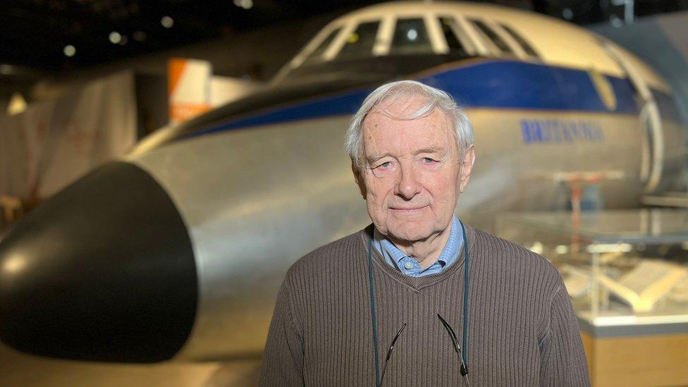 A man in a brown jumper standing in front of a plane