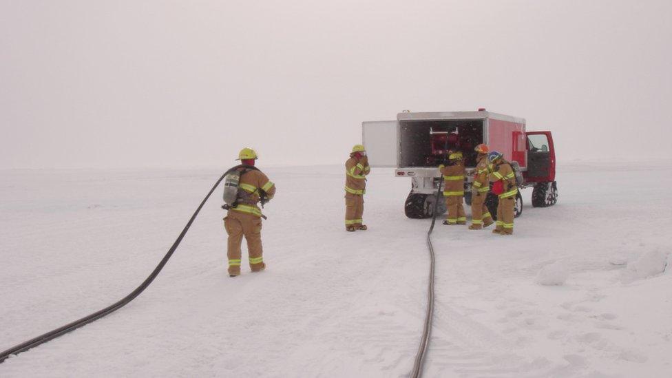 Five fire-fighters work from a vehicle parked on the ice. Hoses stretch behind them and the sky is white.