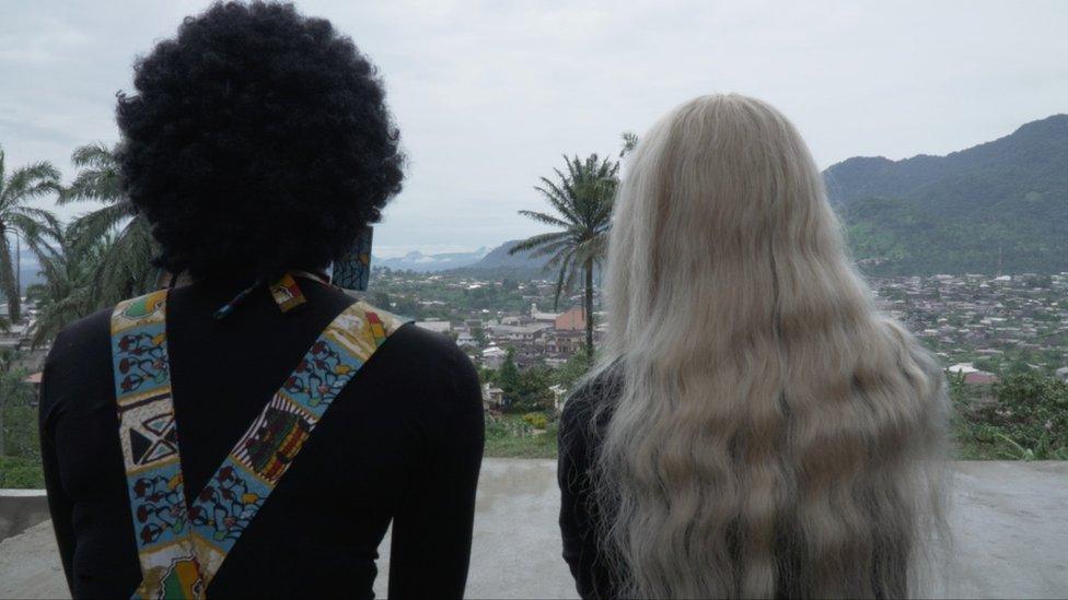 Two women, one with afro, another in a blonde wig, watch the city their backs turned to the camera