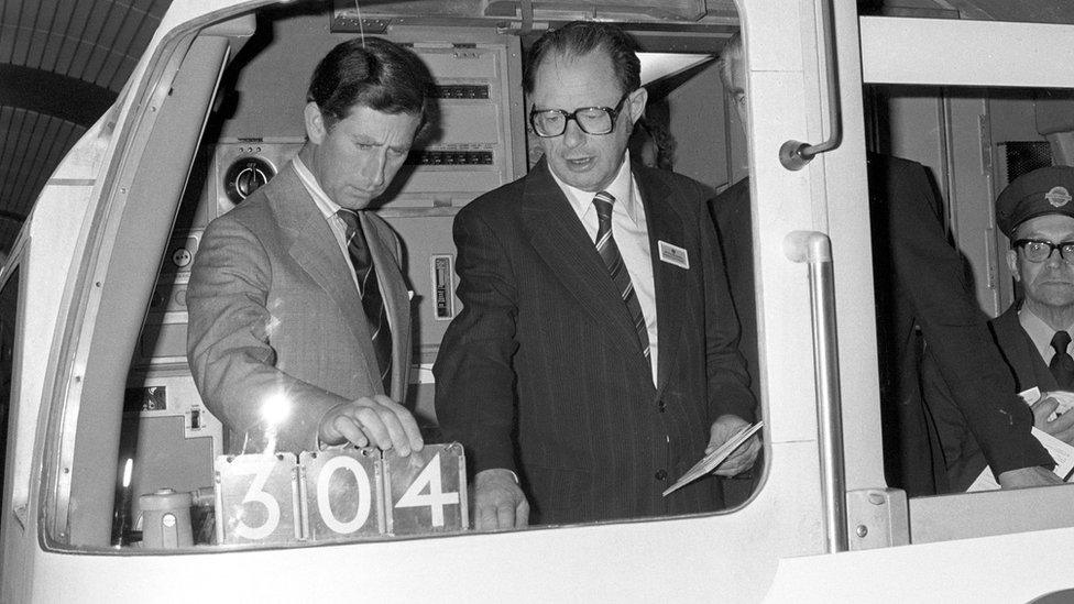 London Transport official showing the Prince Charles, Prince of Wales, the controls in the driver's cab of a Jubilee Line train