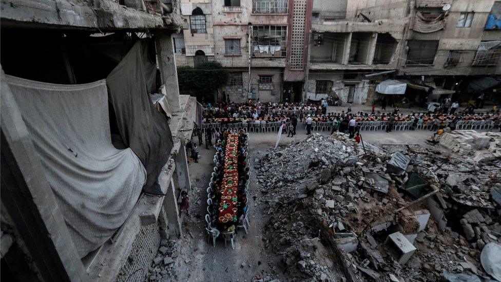 People wait for Iftar, evening meal at the end of daily Ramadan fast at sunset, next to rubble and destroyed houses, which were damaged after air strikes, in Douma, Syria, 17 June 2017.