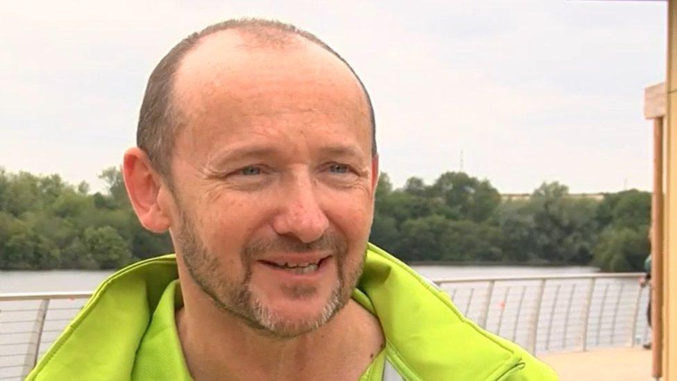 Ian Blackwell with short dark hair and wearing a yellow top stands in front of a lake