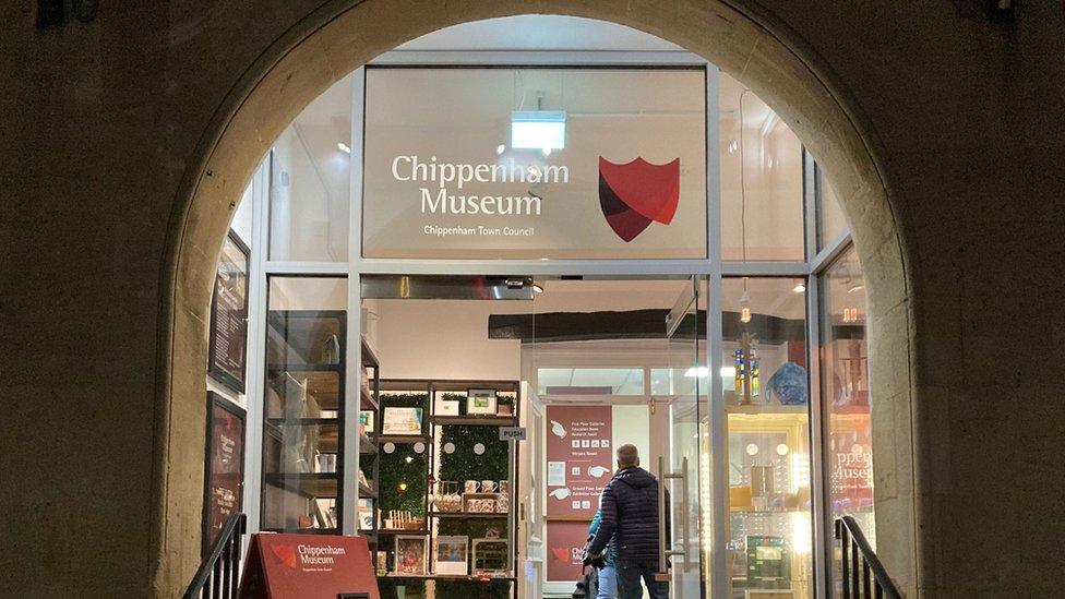 The front of Chippenham museum's entrance showing the lit-up archway and glass door on a dark night