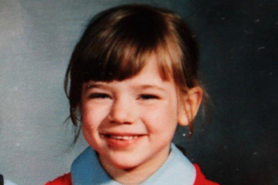 A young girl with brown hair in a red school jumper smiles at the camera