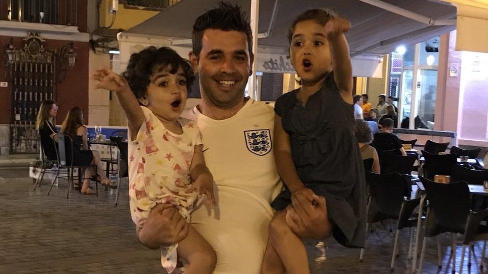 A man in an England shirt holds his two children. Both girls are yelling and gesturing.