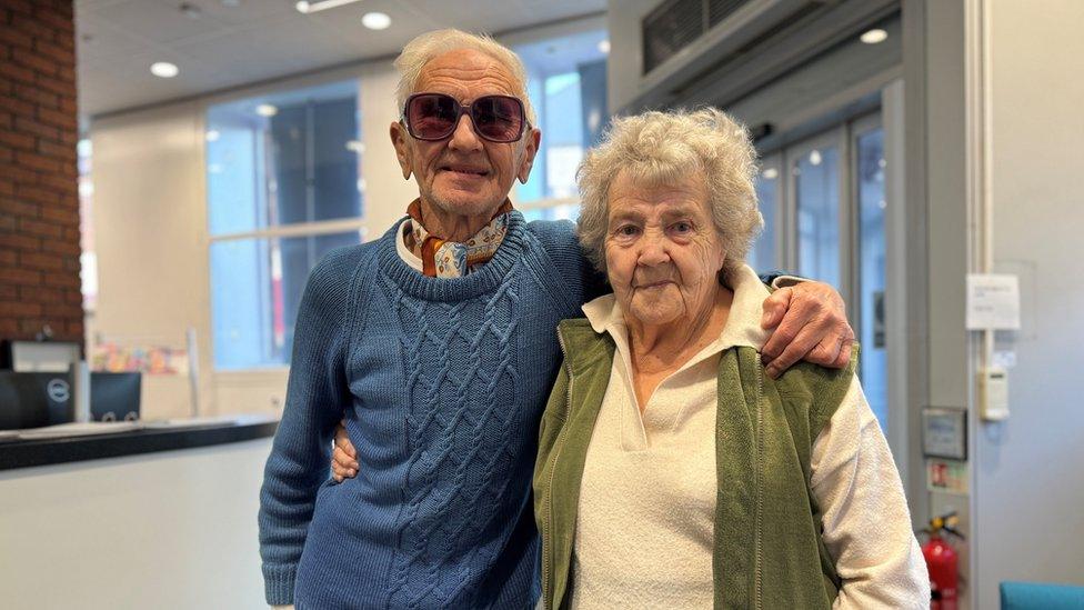 Man with short white hair and dark glasses wearing a blue sweater has his arm round a woman wearing a green gilet over a white shirt