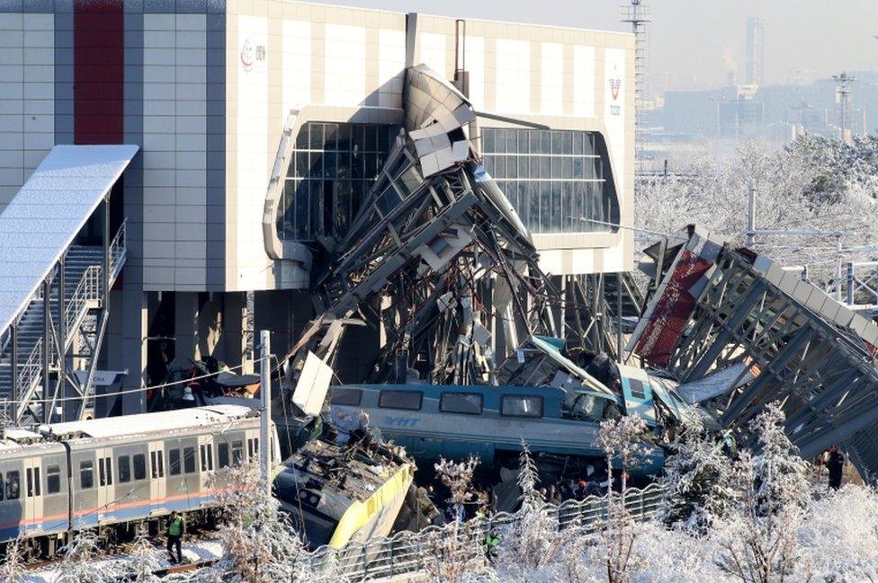 An image shows some train wagons derailed and the metal overpass split in two where the train hit it