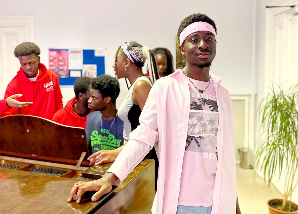 Andrea Menlah from the Reggae Roots choir wearing a pink bandana. He's facing the camera with his fellow choir members in the background.