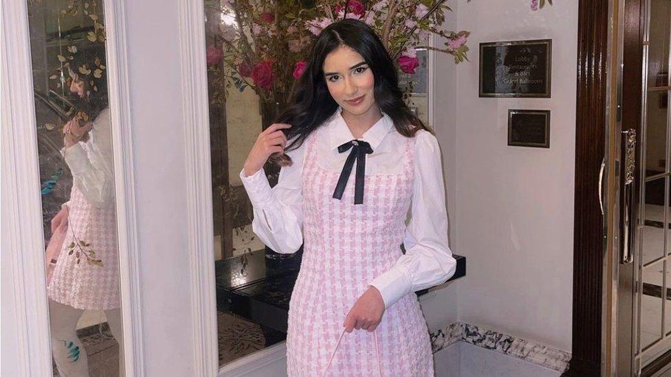 Amira is posing in front of a rose tree which has dark pink flower heads. She is wearing a baby pink and white gingham dress with a white blouse underneath, which has a black bow tied around the collar. Her black hair is down and she is pushing back the right hand side of it with her right hand. Her left hand is holding the strap of a light pink handbag. She is looking directly into the camera and smiling.