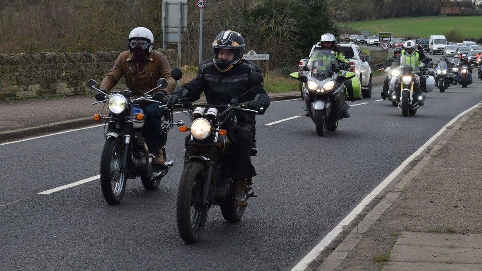 Two bikers lead a procession of motorcycles along a road. Other vehicles are visible in the line.