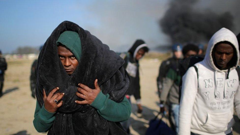 Migrants leave Calais' Jungle camp as smoke billows above the Jungle camp. Photo: 26 October 2016