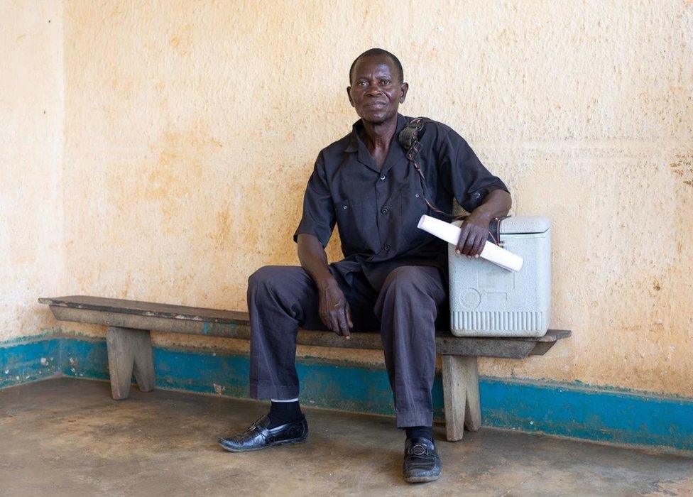 Mulalu Lwesso sits on a wooden bench with his coolbox next to him