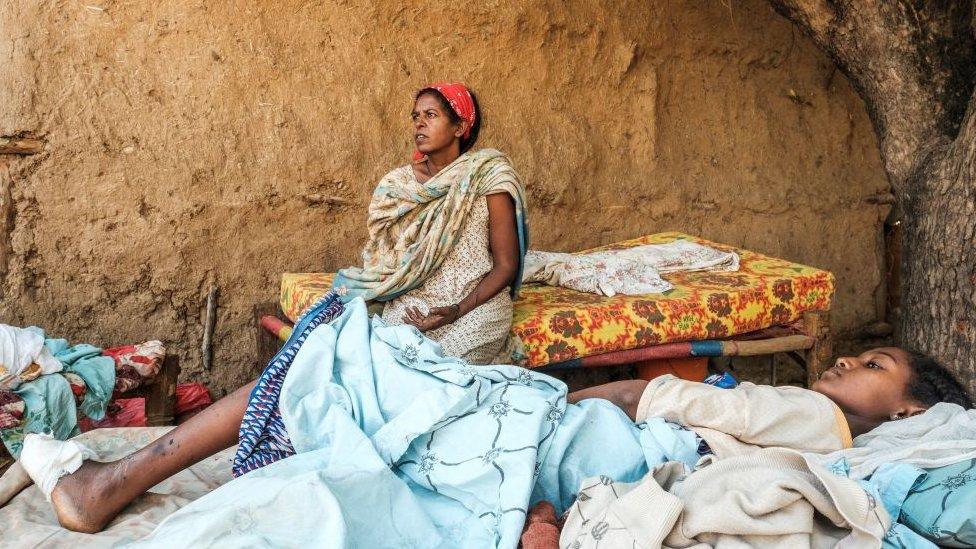 Woman sitting on a bed with someone else lying on a mattress in the foreground