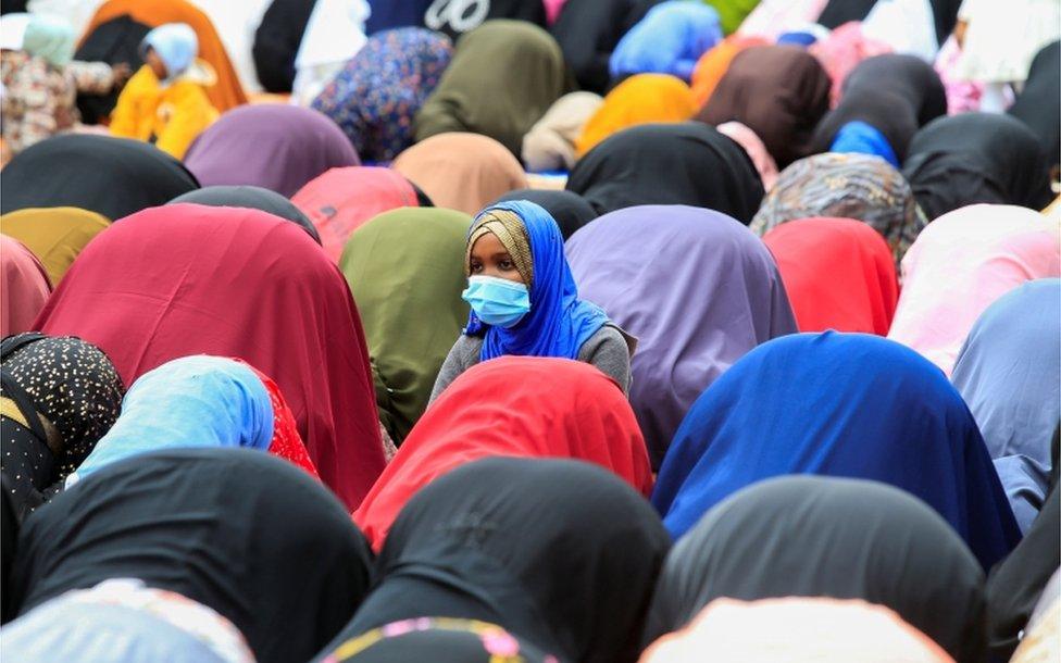 A woman is seen kneeling upright whole others around her are bend forward in prayer.