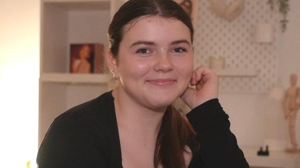 Martha, with her hair tied up and wearing a black top, smiles and looks into the camera