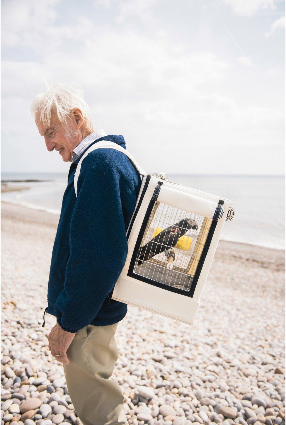 Portrait of Roy on the beach with an Africa grey parrot in a cage on his back