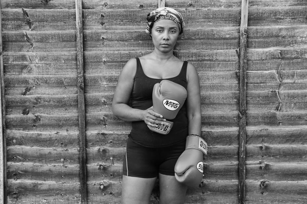 A woman poses against a wooden fence holding boxing gloves