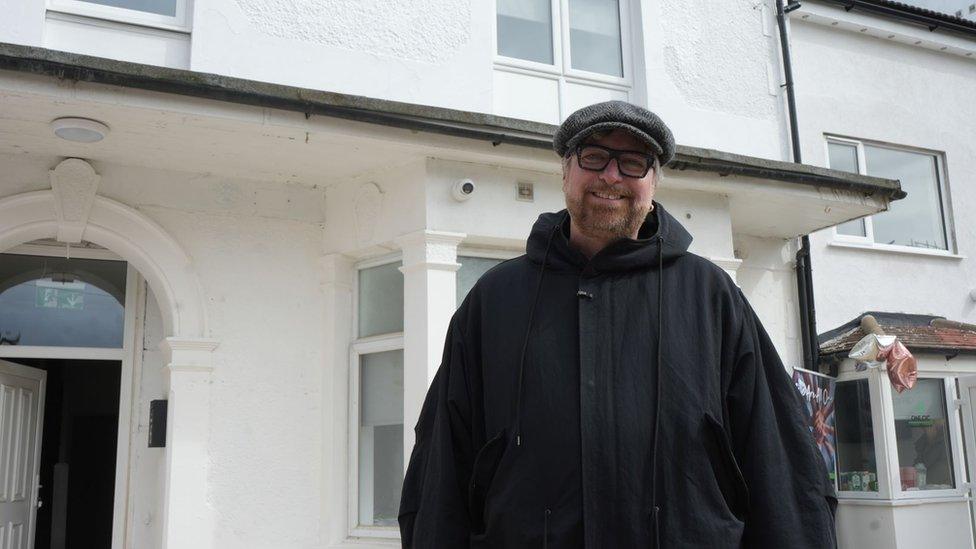 Mark Lovell wearing a cap standing in front of a white building