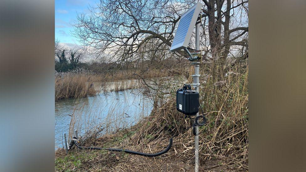 A metal pole stands on the river bank, mounted with a small solar panel and a black box. A flexible black pipe leads into the river to collect samples. The trees are bare and it looks like autumn or winter.