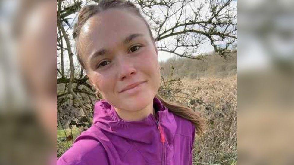 Kit Birks is smiling at the camera, she has brown eyes and her brown hair pulled back into a ponytail. She is wearing a purple waterproof jacket.