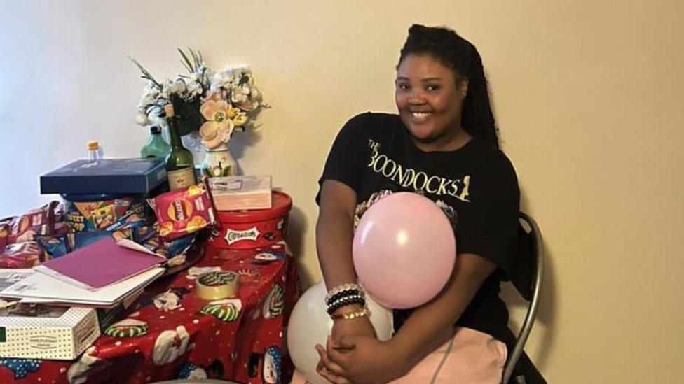 Melissa Zoglie wears a black t-shirt, sitting and holding a pink balloon next a table with gifts on it