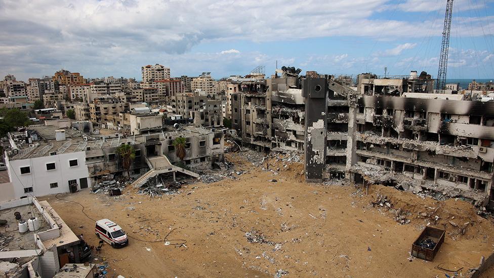 an aerial shot of the ruins of al-shifa hospital taken in september 2024. most of the buildings are in ruins around a barren foreground but part of one building on the left is a bright white and a single white vehicle marked with a red crescent sits in front.