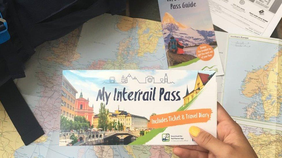 A traveller holds an Interrail pass next to a map of Europe and a backpack