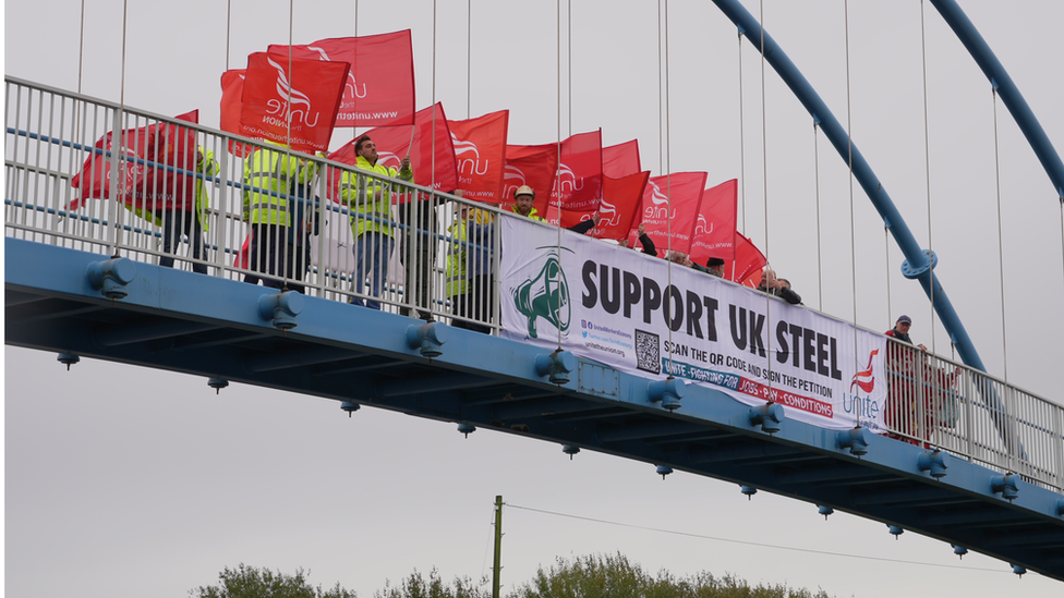 Unite members on bridge waving flags behind sign which reads "support UK steel"