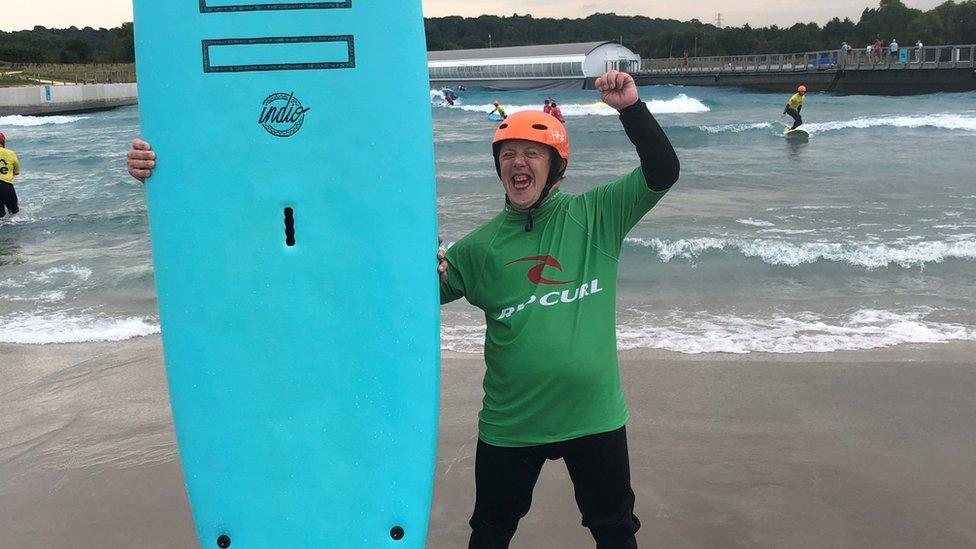 Sam wearing a green wet suit, holding up a surf board