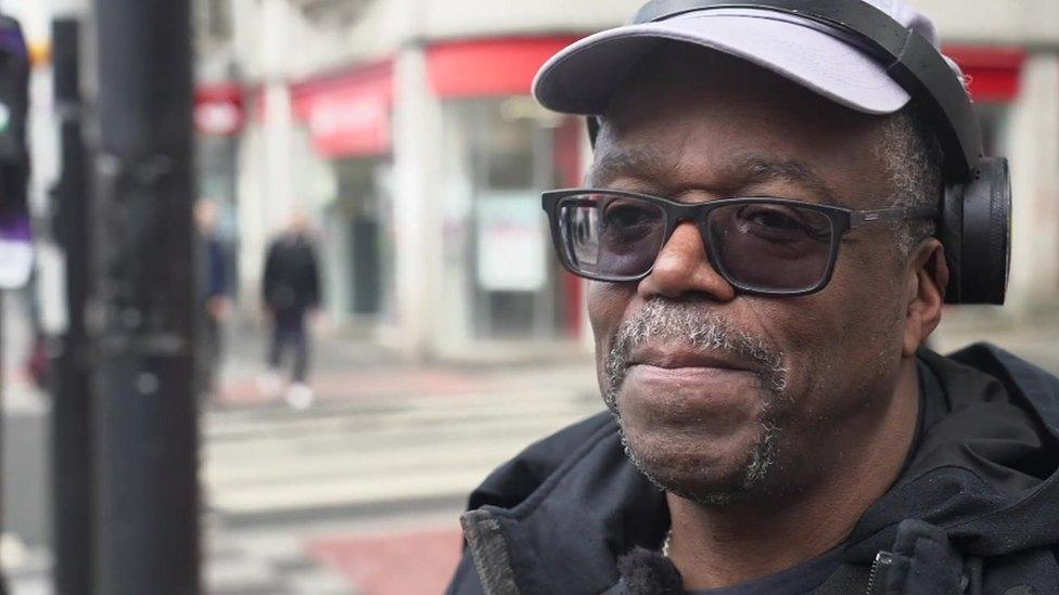 An old man wearing a light hat and headphones standing on a street in Croydon