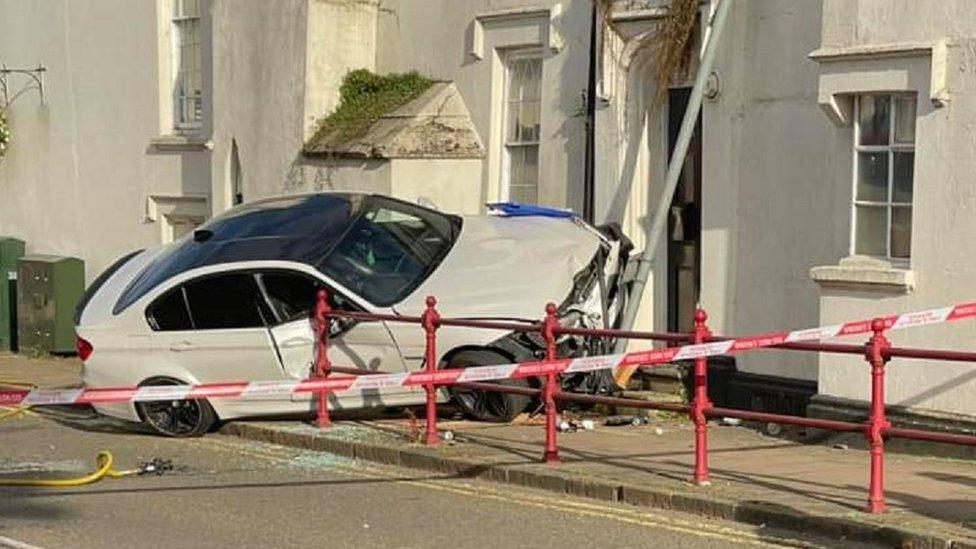 White BMW on pavement with nearside stuck in wall and off the ground. Police tape is visible.