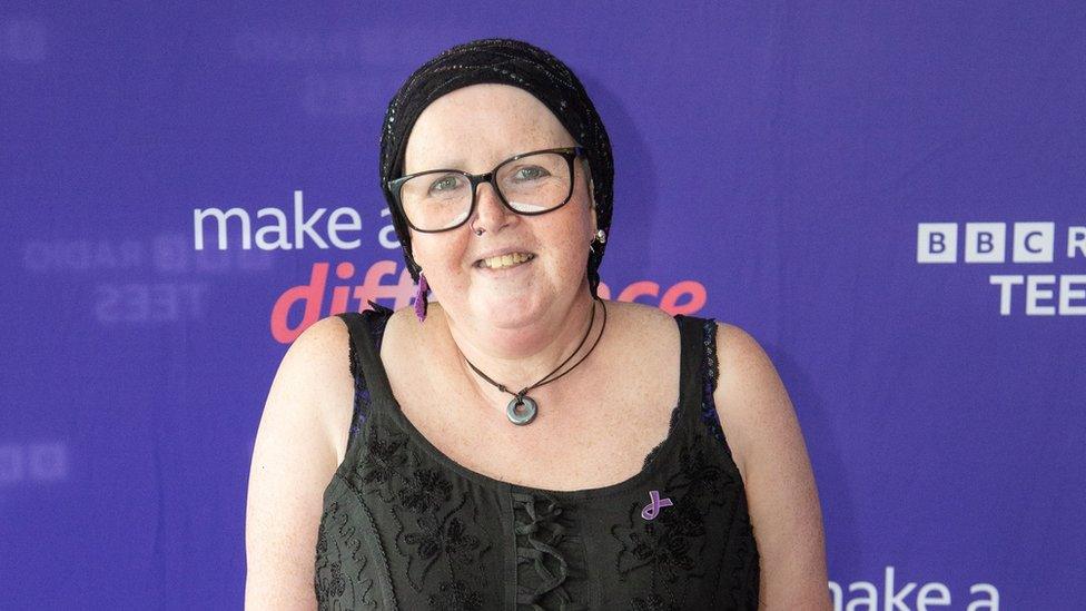 A woman in a black dress, headscarf and glasses smiles at the camera