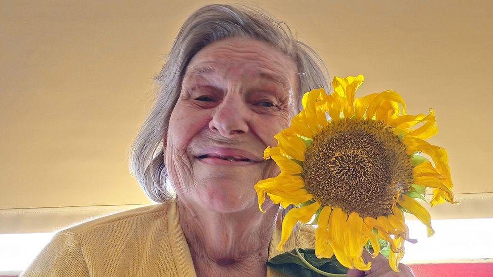 Ann Holmes is 82 and has shoulder length white hair, she is smiling and is holding a sunflower to her left. She is pictured wearing a yellow cardigan and is a room with a mustard coloured wall.