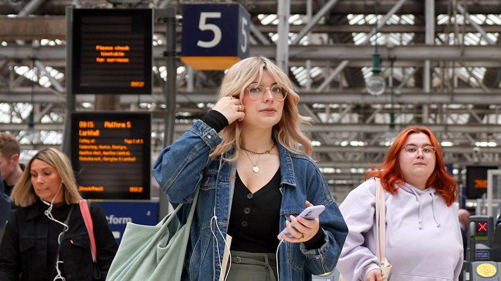 Members of the public make their way through Central Station in Glasgow, Scotland, including a woman with blonde hair, a denim jacket and glasses.