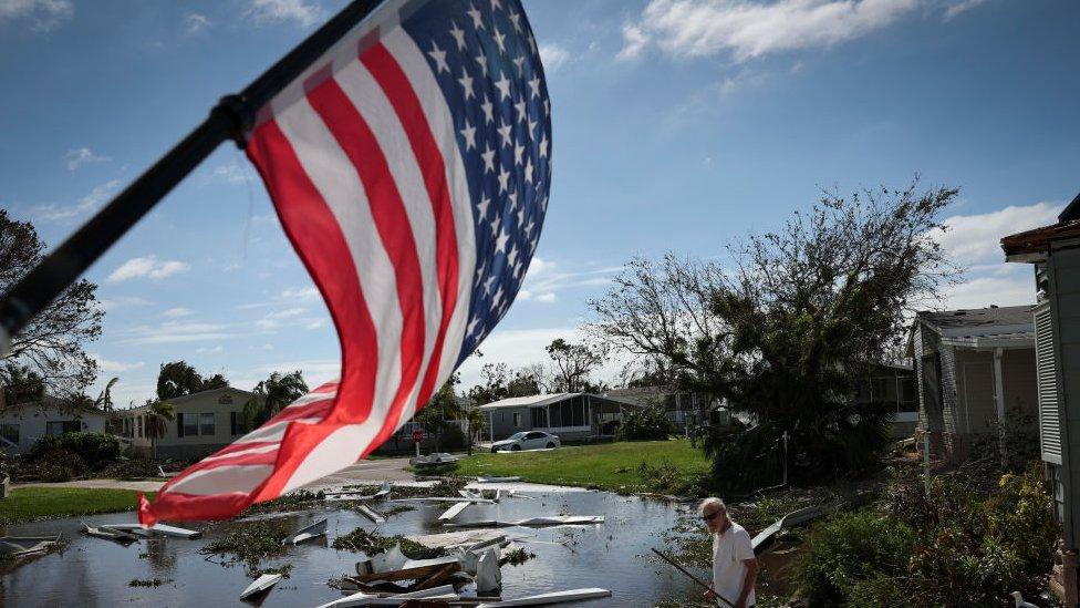 still shows the united states flag waving in the foreground with a florida resident looking over the damage from hurricane ianin the background