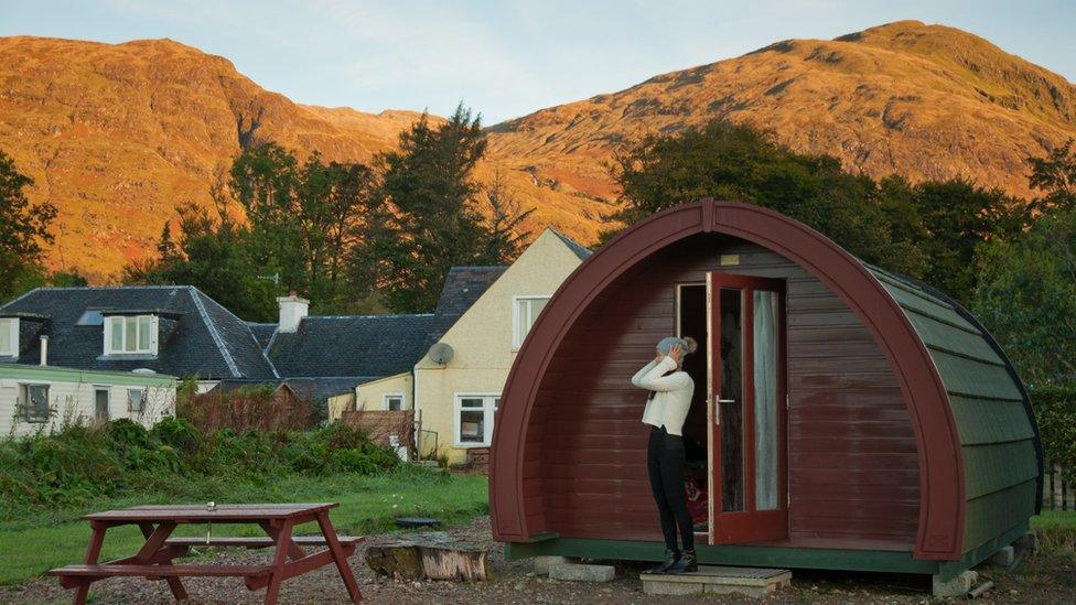 An Asian girl wearing a woolly jumper adjust her woolly hat with her hands while stretching in a cold morning beside a cabin Pod in Clovullin, Fort William, where hills can be seen on the background.