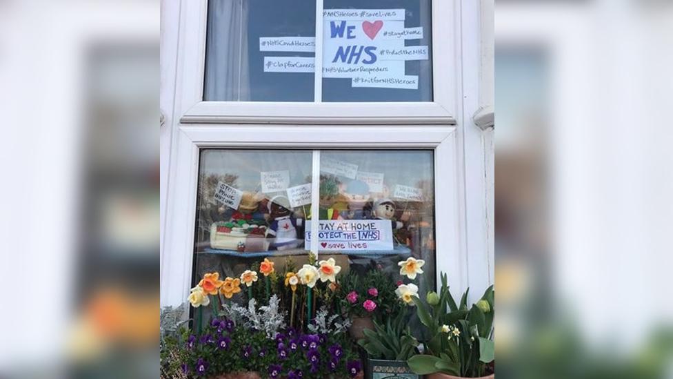 Knitted figures in window with signs "Stay at home. Protect the NHS" and "We stayed at work for you now stay at home for us please"