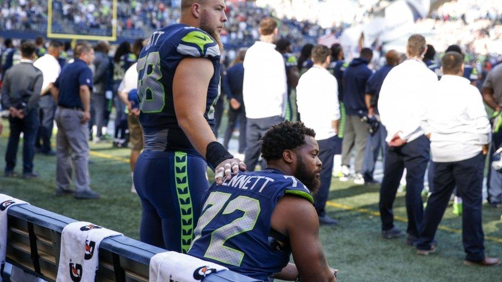 Bennett is pictured sitting amid people standing for anthem