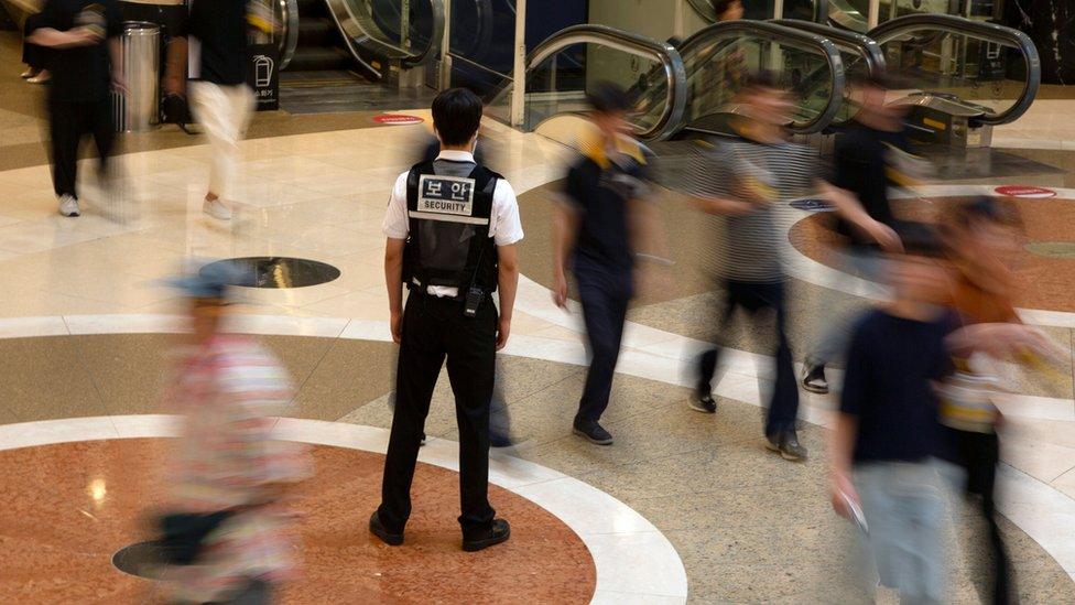 A police officer stands in the middle of passing shoppers at a shopping centre which was the site of a mass stabbing attack on 3 August