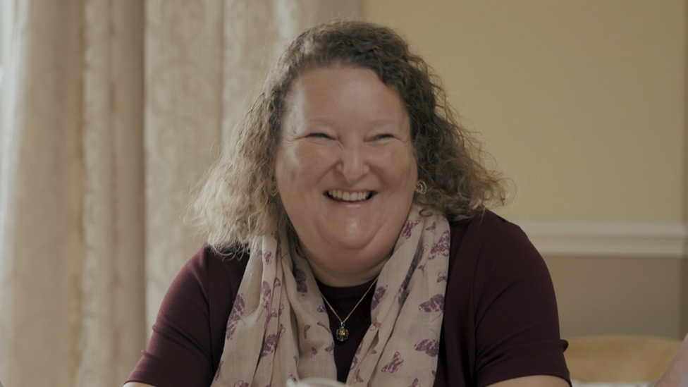 Kerry Smith, who has shoulder-length, wavy hair, and is wearing a scarf and a necklace, is shown in a living room. She is smiling.