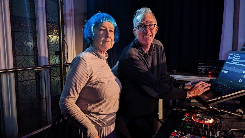 Steve Carpenter next to some DJ equipment with a woman in a blue wig