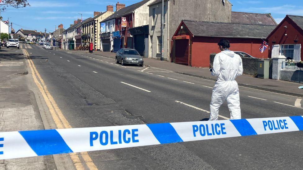 Blue and white police tape across a road, someone in a white forensics suit is walking up the road