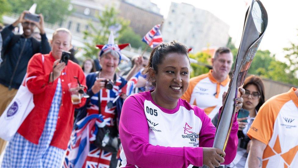 a woman smiles while holding a silver baton while onlookers take pictures