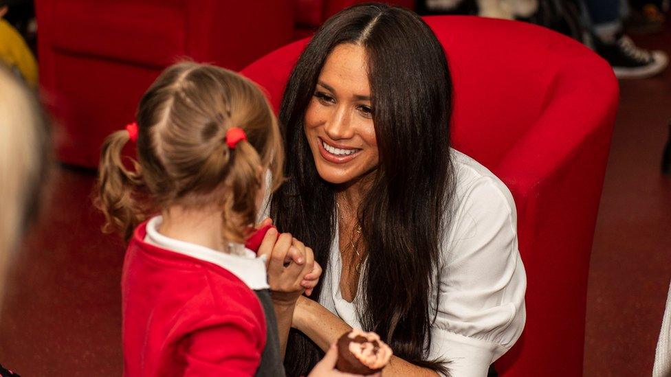 The Duchess of Sussex crouches receives a cake from a little girl at Windsor's Broom Farm Community Centre