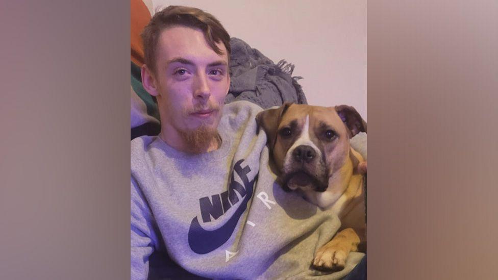 Charlie Stephen David Greaves smiles at the camera while hugging his dog. He has a short beard and moustache and his dog is a tan and white crossbreed. 