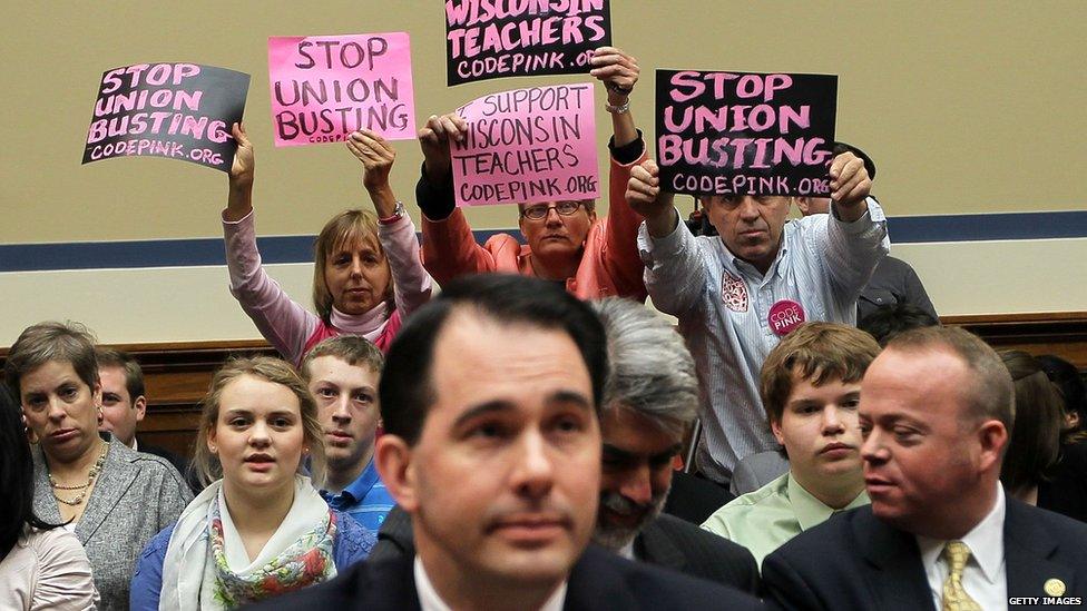 Members of Code Pink (L-R) Medea Benjamin, Liz Hourican and Tighe Barry, hold signs to protest as Wisconsin Gov. Scott Walker (C) takes his seat during a hearing before the House Oversight and Government Reform Committee April 14, 2011 on Capitol Hill in Washington, DC