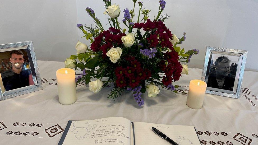 A flower arrangement, lit candles, framed photos of Josh and Gary Dunmore and a book of condolence, with a pen, on a table