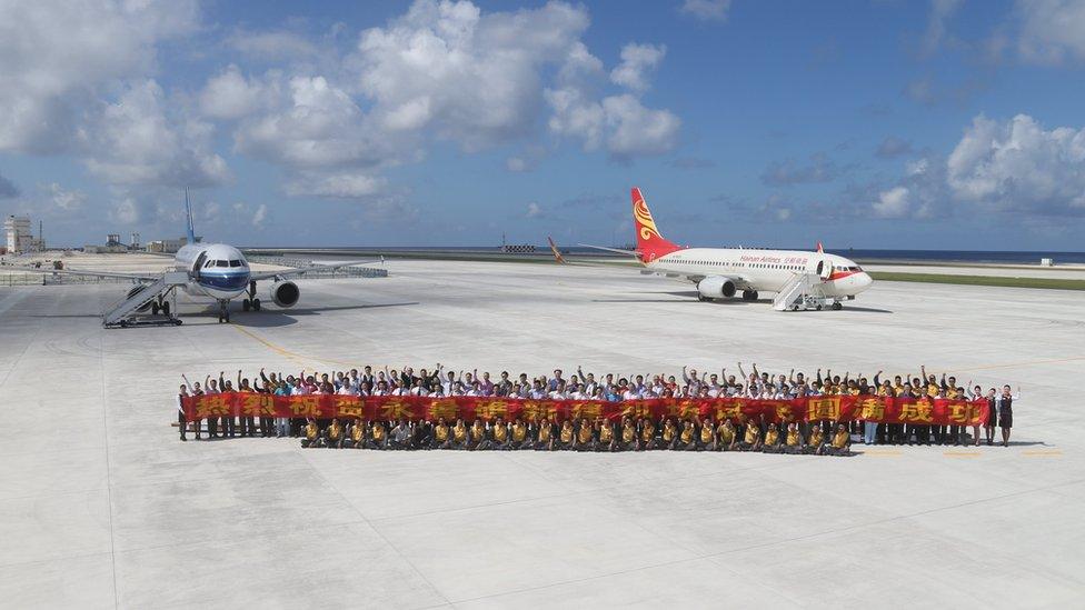 Dozens of people on tarmac of artificial island, with two planes behind, holding up a banner in Chinese